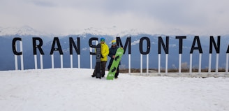 Two people are standing on a snowy mountain landscape, holding snowboards in front of large letters spelling 'Crans Montana'. Snow-covered mountains are visible in the background.