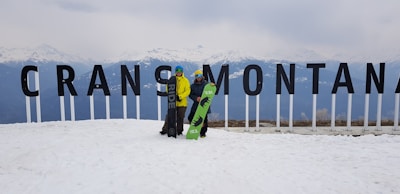 Two people are standing on a snowy mountain landscape, holding snowboards in front of large letters spelling 'Crans Montana'. Snow-covered mountains are visible in the background.