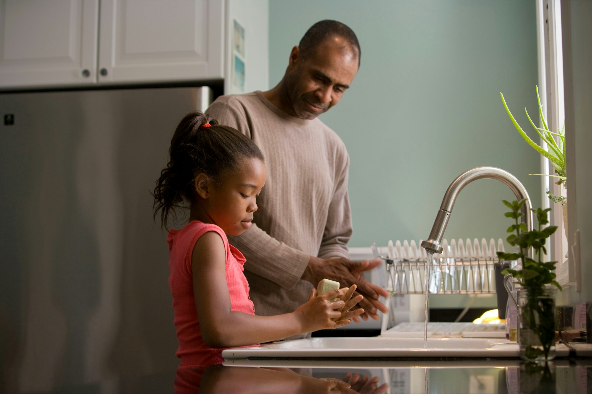This African-American father was shown in the process of teaching his young daughter how to properly wash her hands at their kitchen sink, briskly rubbing her soapy hands together under fresh running tap water, in order to remove germs, and contaminants, thereby, reducing the spread of pathogens, and the ingestion of environmental chemicals or toxins. Children are taught to recite the Happy Birthday song, during hand washing, allotting enough time to completely clean their hands.
