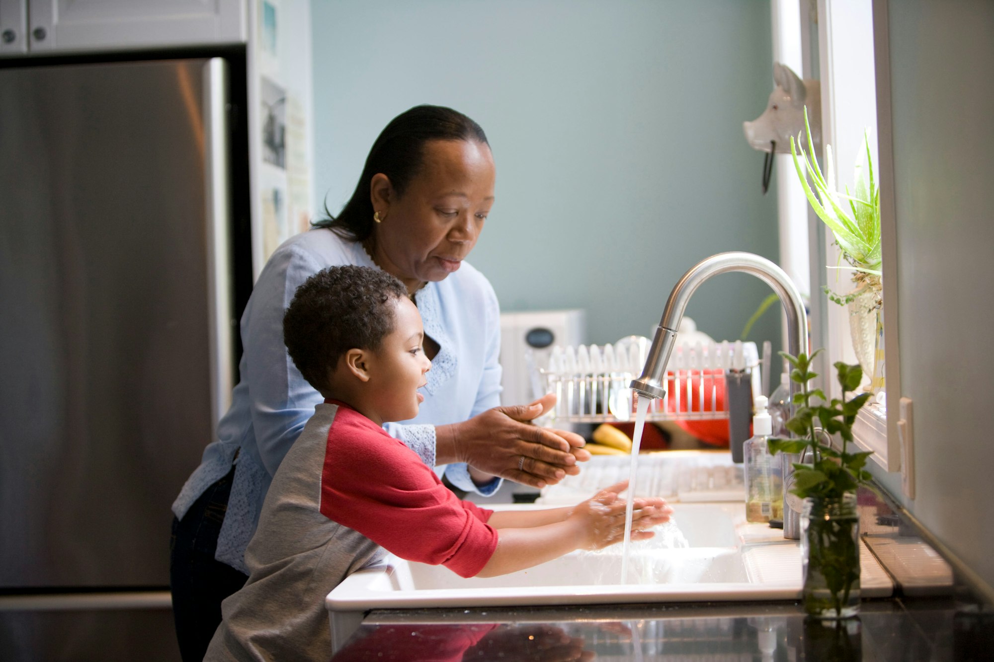 This African-American mother was shown in the process of teaching her young son how to properly wash his hands at their kitchen sink, briskly rubbing his soapy hands together under fresh running tap water, in order to remove germs, and contaminants, thereby, reducing the spread of pathogens, and the ingestion of environmental chemicals or toxins. Children are taught to recite the Happy Birthday song, during hand washing, allotting enough time to completely clean their hands.