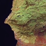 A satellite image that depicts a vast expanse of rugged terrain with visible mountains and valleys. The landscape reveals varying elevations with lush greenery concentrated in some areas, indicating vegetation, while other sections are more barren. The left side is bordered by a dark space representing a large body of water.