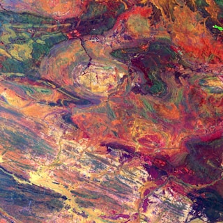 A vibrant satellite image showcasing a geological formation with swirling patterns. The landscape features a mix of rugged textures and undulating contours, with colors ranging from deep reds and purples to bright yellows and greens. The central area appears to be a circular crater-like structure.
