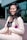 A woman is smiling and posing in a light-colored suit with a black lace top. She has long, wavy black hair and is standing in front of a modern glass building. A purple flower with green leaves is pinned to her lapel, adding a pop of color.