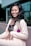 A woman is smiling and posing in a light-colored suit with a black lace top. She has long, wavy black hair and is standing in front of a modern glass building. A purple flower with green leaves is pinned to her lapel, adding a pop of color.