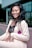 A woman is smiling and posing in a light-colored suit with a black lace top. She has long, wavy black hair and is standing in front of a modern glass building. A purple flower with green leaves is pinned to her lapel, adding a pop of color.