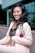 A woman is smiling and posing in a light-colored suit with a black lace top. She has long, wavy black hair and is standing in front of a modern glass building. A purple flower with green leaves is pinned to her lapel, adding a pop of color.