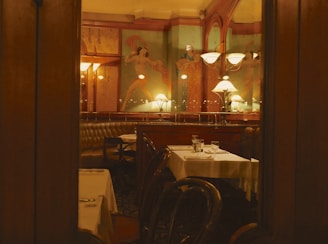 A cozy restaurant interior featuring dim lighting with elegantly set tables and cushioned seating. The walls have ornate decorations, including large paintings or murals with dancers. There are classic wooden chairs and stylish lamps illuminating the space, creating a warm and inviting atmosphere.