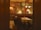 A cozy restaurant interior featuring dim lighting with elegantly set tables and cushioned seating. The walls have ornate decorations, including large paintings or murals with dancers. There are classic wooden chairs and stylish lamps illuminating the space, creating a warm and inviting atmosphere.