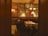 A cozy restaurant interior featuring dim lighting with elegantly set tables and cushioned seating. The walls have ornate decorations, including large paintings or murals with dancers. There are classic wooden chairs and stylish lamps illuminating the space, creating a warm and inviting atmosphere.
