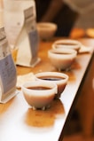 Several small cups filled with coffee are arranged in a line on a wooden table. In the background, there are upright coffee bags blurred and other objects slightly out of focus.