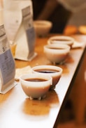 Several small cups filled with coffee are arranged in a line on a wooden table. In the background, there are upright coffee bags blurred and other objects slightly out of focus.