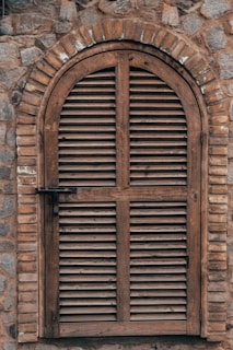 An arched wooden shutter window is set into a textured stone wall. The wood has a weathered appearance, with horizontal slats and a simple latch on the left side. The stone wall is comprised of irregular stones in various shades of gray and brown, contributing to a rustic aesthetic.