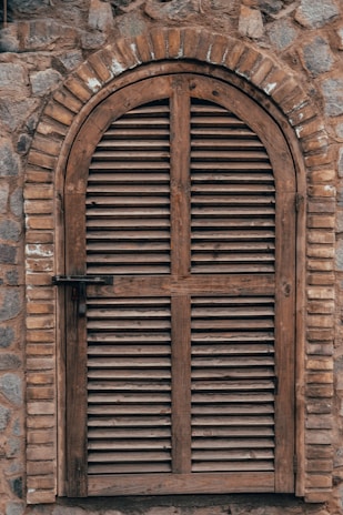 An arched wooden shutter window is set into a textured stone wall. The wood has a weathered appearance, with horizontal slats and a simple latch on the left side. The stone wall is comprised of irregular stones in various shades of gray and brown, contributing to a rustic aesthetic.