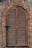 An arched wooden shutter window is set into a textured stone wall. The wood has a weathered appearance, with horizontal slats and a simple latch on the left side. The stone wall is comprised of irregular stones in various shades of gray and brown, contributing to a rustic aesthetic.