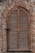 An arched wooden shutter window is set into a textured stone wall. The wood has a weathered appearance, with horizontal slats and a simple latch on the left side. The stone wall is comprised of irregular stones in various shades of gray and brown, contributing to a rustic aesthetic.