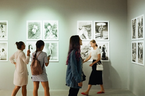 Several people walk and observe black-and-white photographs displayed on a gallery wall. The visitors appear engaged with the art, with some standing while others move through the space.