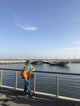 A person wearing a teal headscarf and yellow top stands on a waterfront promenade, leaning casually against a metal railing. They are holding an orange folder and looking upwards. The background features a calm body of water, some industrial structures, a boat, and a breakwater with rocks under a clear blue sky.