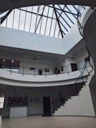 A spacious interior with a modern design featuring a large skylight with black metal framing. The space includes a two-level structure with curved balconies and black railings, surrounded by white walls. Several potted plants adorn the upper level, and a staircase leads up to the higher floor.