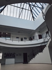 A spacious interior with a modern design featuring a large skylight with black metal framing. The space includes a two-level structure with curved balconies and black railings, surrounded by white walls. Several potted plants adorn the upper level, and a staircase leads up to the higher floor.