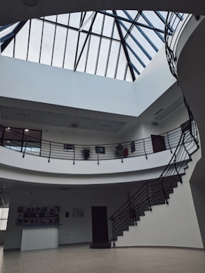 A spacious interior with a modern design featuring a large skylight with black metal framing. The space includes a two-level structure with curved balconies and black railings, surrounded by white walls. Several potted plants adorn the upper level, and a staircase leads up to the higher floor.