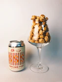 A can of 'Popcorn Pimp' sweet and salty imperial stout is placed next to a wine glass filled with caramel popcorn. The setup is on a plain, neutral-colored surface with a soft background, creating a simple yet stylish presentation.
