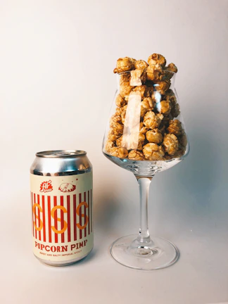 A can of 'Popcorn Pimp' sweet and salty imperial stout is placed next to a wine glass filled with caramel popcorn. The setup is on a plain, neutral-colored surface with a soft background, creating a simple yet stylish presentation.