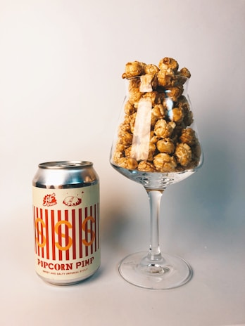 A can of 'Popcorn Pimp' sweet and salty imperial stout is placed next to a wine glass filled with caramel popcorn. The setup is on a plain, neutral-colored surface with a soft background, creating a simple yet stylish presentation.