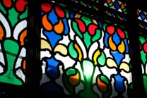 Intricate stained glass with vibrant colors and floral motifs. The design features harmonious arrangements of red, blue, green, yellow, and orange pieces, creating a dazzling effect as light shines through.