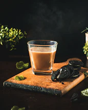 A glass filled with a hot beverage is positioned on a wooden board alongside several chocolate sandwich cookies. Steam rises from the drink, suggesting it is freshly made. Green leaves are scattered around the board, adding a touch of freshness. The background is dark, highlighting the main subjects, with subtle hints of greenery.