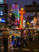 A bustling street scene at night features vibrant neon signs and lights illuminating the area. People are walking along the street, and a tuk-tuk taxi is parked with a driver inside. The atmosphere is lively and colorful.