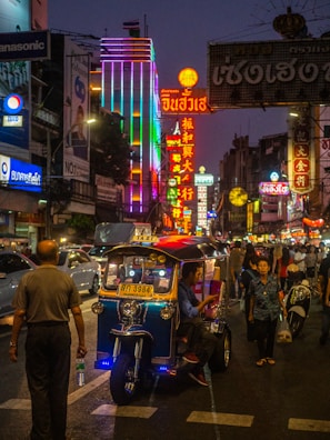 A bustling street scene at night features vibrant neon signs and lights illuminating the area. People are walking along the street, and a tuk-tuk taxi is parked with a driver inside. The atmosphere is lively and colorful.