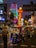 A bustling street scene at night features vibrant neon signs and lights illuminating the area. People are walking along the street, and a tuk-tuk taxi is parked with a driver inside. The atmosphere is lively and colorful.