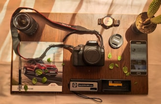 A wooden table holds a variety of photography-related items neatly arranged. A Canon EOS 2000D camera sits at the center, accompanied by a lens and lens cap. To the left, a magazine features an image of a red SUV. Nearby, a wristwatch, potted cactus, and Samsung smartphone with an Instagram feed are visible. Scattered green leaves add a touch of nature to the composition.