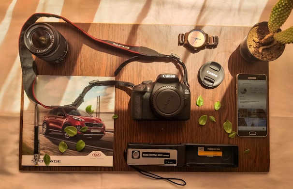 A wooden table holds a variety of photography-related items neatly arranged. A Canon EOS 2000D camera sits at the center, accompanied by a lens and lens cap. To the left, a magazine features an image of a red SUV. Nearby, a wristwatch, potted cactus, and Samsung smartphone with an Instagram feed are visible. Scattered green leaves add a touch of nature to the composition.