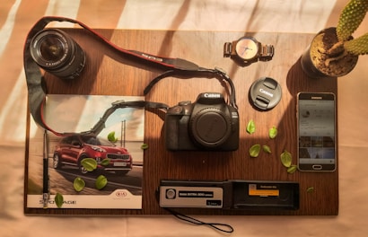 A wooden table holds a variety of photography-related items neatly arranged. A Canon EOS 2000D camera sits at the center, accompanied by a lens and lens cap. To the left, a magazine features an image of a red SUV. Nearby, a wristwatch, potted cactus, and Samsung smartphone with an Instagram feed are visible. Scattered green leaves add a touch of nature to the composition.