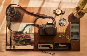 A wooden table holds a variety of photography-related items neatly arranged. A Canon EOS 2000D camera sits at the center, accompanied by a lens and lens cap. To the left, a magazine features an image of a red SUV. Nearby, a wristwatch, potted cactus, and Samsung smartphone with an Instagram feed are visible. Scattered green leaves add a touch of nature to the composition.