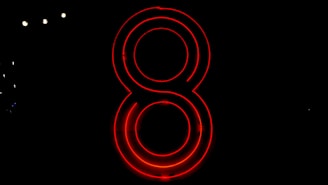 A neon red number eight glows brightly against a completely black background. The light reflects off its circular outlines, creating a striking visual contrast.