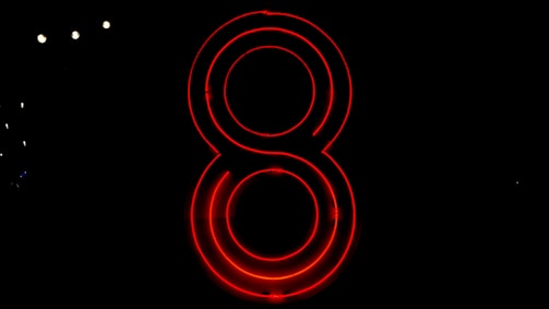 A neon red number eight glows brightly against a completely black background. The light reflects off its circular outlines, creating a striking visual contrast.