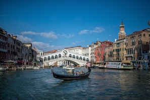 A gondola navigates the waters of a canal in Venice, with the iconic Rialto Bridge in the background. The scene is framed by historic buildings in various shades of red, yellow, and beige, with a clear blue sky above.