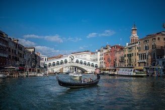 A gondola navigates the waters of a canal in Venice, with the iconic Rialto Bridge in the background. The scene is framed by historic buildings in various shades of red, yellow, and beige, with a clear blue sky above.