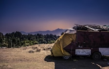 A rustic, makeshift shelter is parked on a dirt ground, covered partially with a yellow tarpaulin. Nearby, a sign indicates the acceptance of digital payments through Paytm. The backdrop features a serene landscape with distant mountains under a clear, darkening sky.