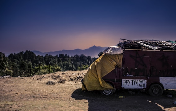 A rustic, makeshift shelter is parked on a dirt ground, covered partially with a yellow tarpaulin. Nearby, a sign indicates the acceptance of digital payments through Paytm. The backdrop features a serene landscape with distant mountains under a clear, darkening sky.