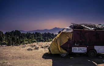 A rustic, makeshift shelter is parked on a dirt ground, covered partially with a yellow tarpaulin. Nearby, a sign indicates the acceptance of digital payments through Paytm. The backdrop features a serene landscape with distant mountains under a clear, darkening sky.