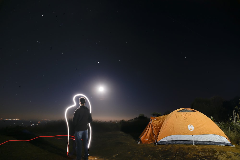 man in black jacket standing near orange dome tent during night time