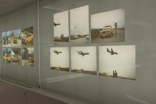 A series of framed photographs displayed on a wall in a grid pattern. The images feature a vintage orange car and scenes of airplanes flying overhead. Some photos show people around the car, with a scenic outdoor setting in the background.