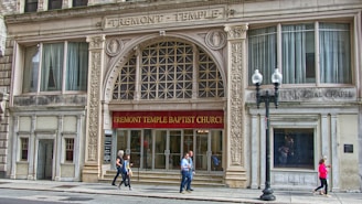 The facade of a historical building with ornate architectural details, featuring the Tremont Temple Baptist Church. The entrance is marked by a large arch with decorative elements and a red banner displaying the church's name. Pedestrians are walking along the sidewalk in front of the church.
