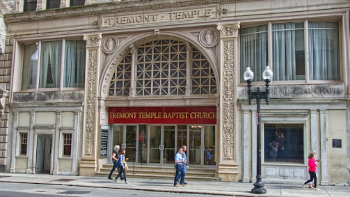 The facade of a historical building with ornate architectural details, featuring the Tremont Temple Baptist Church. The entrance is marked by a large arch with decorative elements and a red banner displaying the church's name. Pedestrians are walking along the sidewalk in front of the church.