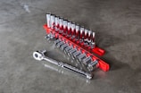 A set of shiny metallic socket wrenches neatly organized on a bright red tool holder placed on a smooth gray concrete surface. The wrenches vary in size, arranged in ascending order with a ratchet handle lying adjacent to them.