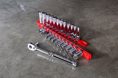 A set of shiny metallic socket wrenches neatly organized on a bright red tool holder placed on a smooth gray concrete surface. The wrenches vary in size, arranged in ascending order with a ratchet handle lying adjacent to them.