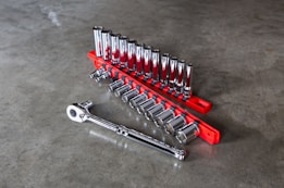 A set of shiny metallic socket wrenches neatly organized on a bright red tool holder placed on a smooth gray concrete surface. The wrenches vary in size, arranged in ascending order with a ratchet handle lying adjacent to them.